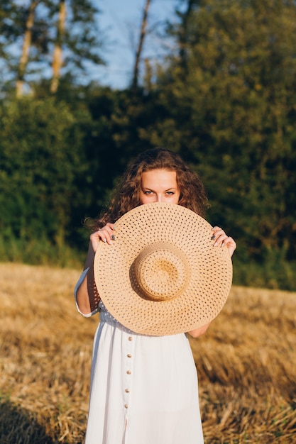 Jovem mulher bonita com cabelo longo encaracolado posa num campo de trigo no verão ao pôr do sol