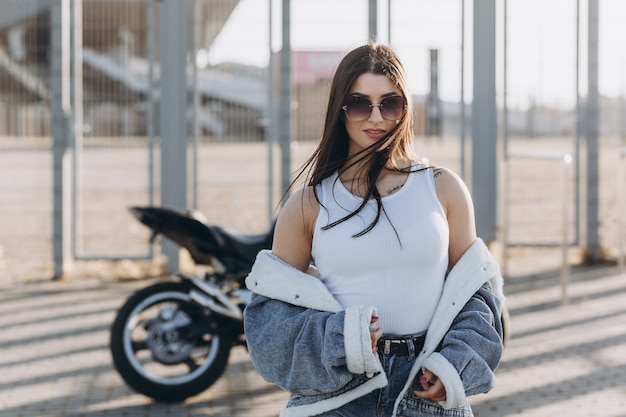 Jovem mulher bonita com cabelo preto, calça jeans e uma jaqueta perto de uma motocicleta esportiva preta