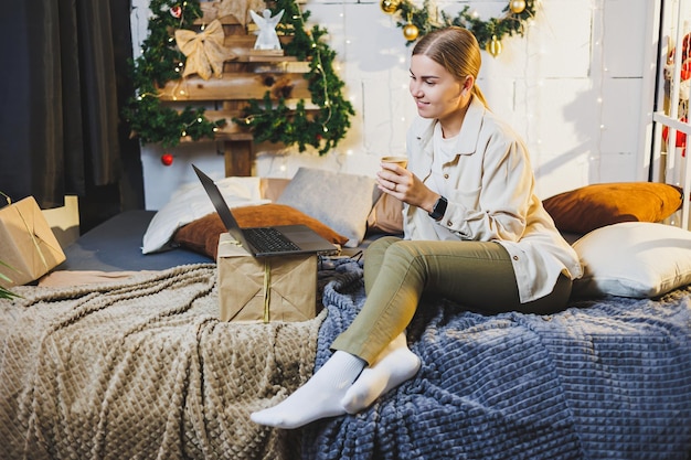 Jovem mulher bonita com cabelos longos loiros em roupas aconchegantes com café nas mãos usando laptop assistindo filme e sorrindo na cama no quarto com árvore de Natal em casa feriados de ano novo