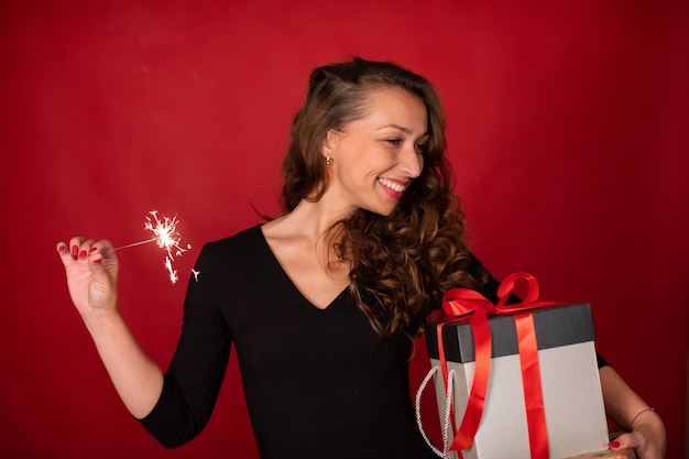 Jovem mulher bonita com caixa de presente e diamante aceso sorrindo sobre fundo vermelho. Festa de Natal, celebração de Réveillon, ideias românticas para presentear, conceito de self-gifter. Copie o espaço
