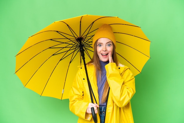 Jovem mulher bonita com casaco à prova de chuva e guarda-chuva sobre fundo croma isolado com surpresa e expressão facial chocada