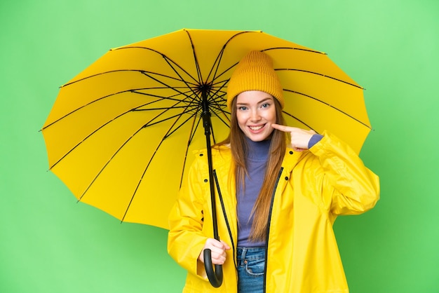 Jovem mulher bonita com casaco à prova de chuva e guarda-chuva sobre fundo croma isolado dando um polegar para cima gesto