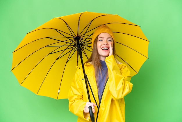 Jovem mulher bonita com casaco à prova de chuva e guarda-chuva sobre fundo croma isolado gritando com a boca aberta