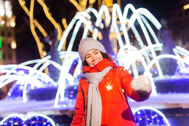 Jovem mulher bonita com chapéu de malha e lenço em pé na cidade com luz de Bengala, diamante. Celebração do conceito e Natal.