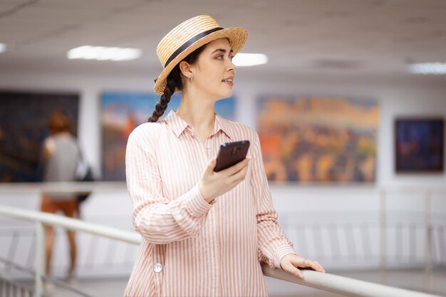 Jovem mulher bonita com chapéu de palha está segurando um smartphone No fundo é a sala de exposições de arte da galeria com pinturas Conceito de educação cultural e tecnologia moderna