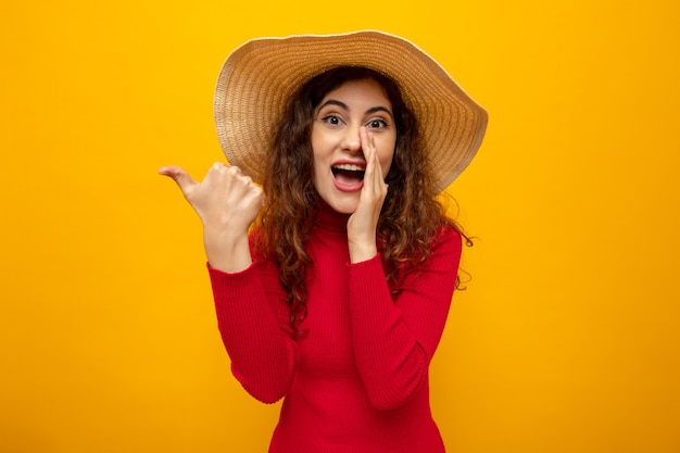 Jovem mulher bonita com gola alta vermelha e chapéu de verão olhando sorrindo, contando um segredo com a mão na boca apontando com o polegar para o lado