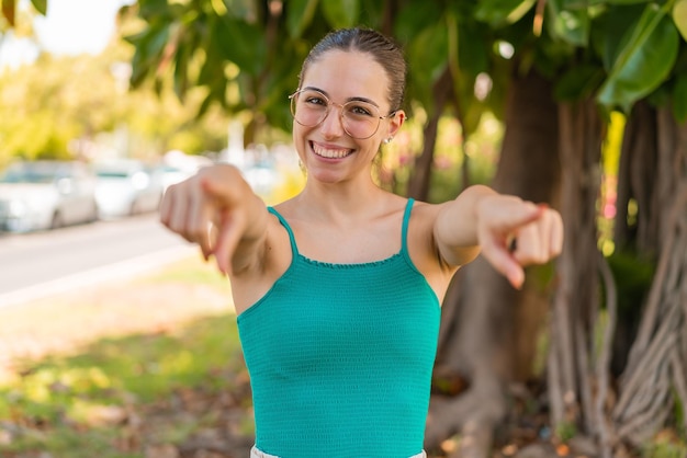 Foto jovem mulher bonita com óculos ao ar livre aponta o dedo para você enquanto sorri