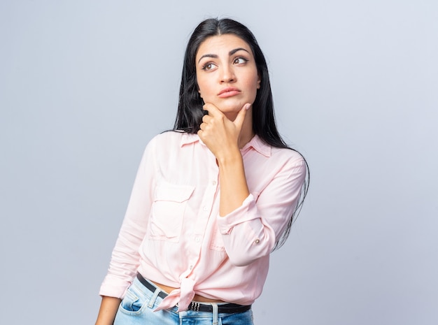 Jovem mulher bonita com roupas casuais olhando para o lado perplexa com a mão no queixo de pé sobre uma parede branca