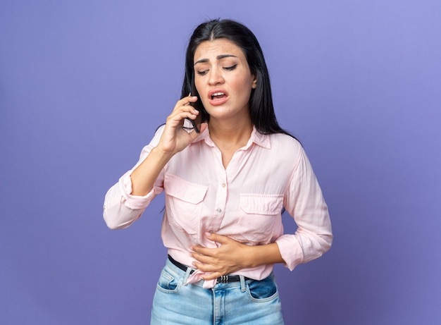 Jovem mulher bonita com roupas casuais parecendo confusa enquanto fala no celular em pé no azul