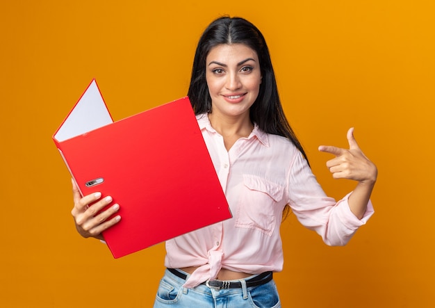 Jovem mulher bonita com roupas casuais segurando uma pasta feliz e satisfeita, sorrindo confiante apontando com o dedo indicador para a pasta em pé sobre a parede laranja