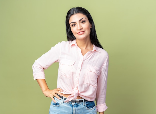 Jovem mulher bonita com roupas casuais, sorrindo, confiante, feliz e positiva em pé sobre a parede verde