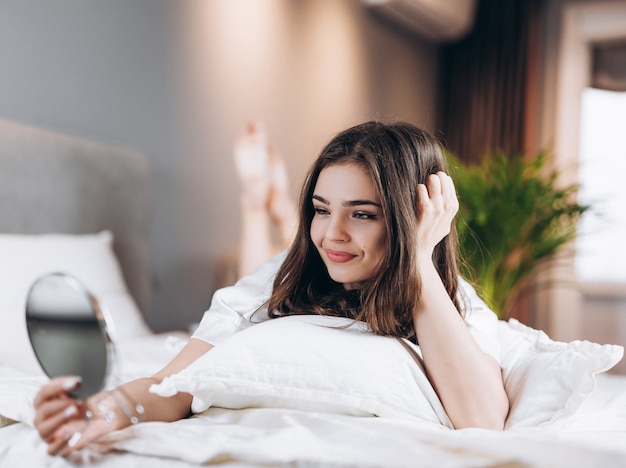 Jovem mulher bonita com um espelho nas mãos no quarto dela. Modelo feminino se olhando no espelho enquanto estava deitado na cama.