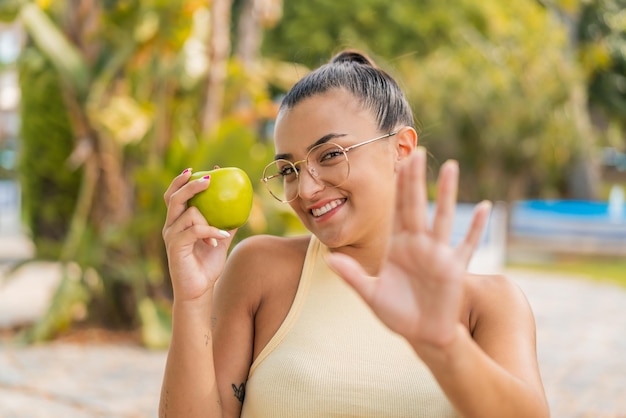 Foto jovem mulher bonita com uma maçã ao ar livre cumprimentando com a mão com expressão feliz