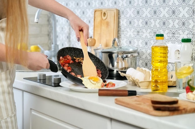 Jovem mulher bonita cozinhando em casa na cozinha, ovos fritos com cogumelos de salsichas e páprica na frigideira. Comida saudável fresca caseira, hobbies e lazer