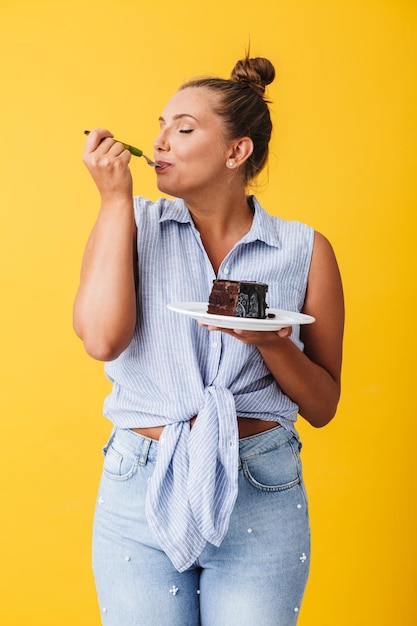 Foto jovem mulher bonita de camisa comendo alegremente bolo de chocolate sobre fundo amarelo isolado