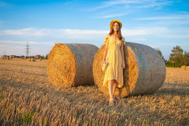 Jovem mulher bonita de vestido amarelo e chapéu perto do fundo do campo com palheiros na hora do pôr do sol