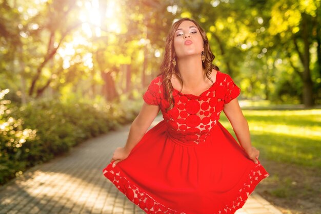 Jovem mulher bonita de vestido vermelho aproveitar o sol no parque. Ela está mandando um beijo e olhando para a câmera.