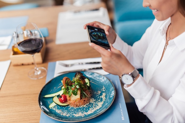 Jovem mulher bonita desfrutando de refeição saborosa e bem decorada. Ela está sentada em um restaurante caro e usando seu smartphone para fotografar alimentos.