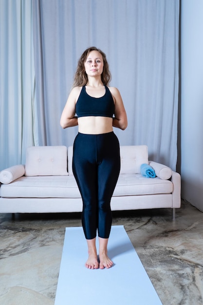 Jovem mulher bonita e saudável em top esportivo e leggings praticando ioga em casa em pose no tapete de ioga