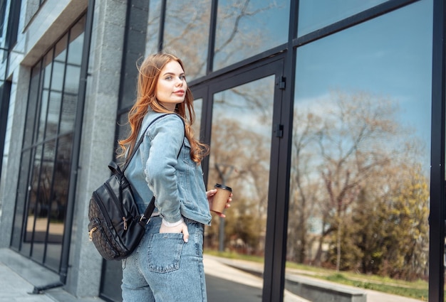 Jovem mulher bonita em jaqueta jeans e segurando uma xícara de café perto das janelas do centro de negócios