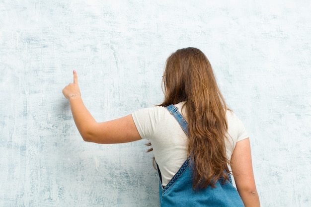 Jovem mulher bonita em pé e apontando para objetar no espaço da cópia, vista traseira contra a parede do grunge