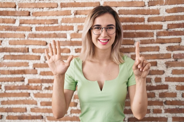 Jovem mulher bonita em pé sobre a parede de tijolos mostrando e apontando para cima com os dedos número seis enquanto sorria confiante e feliz