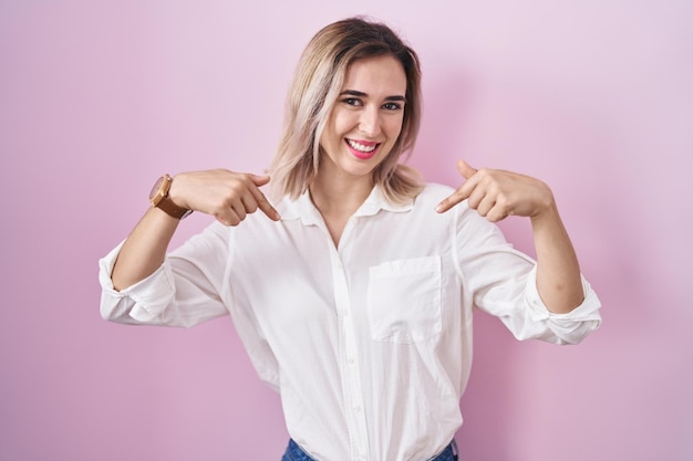 Jovem mulher bonita em pé sobre fundo rosa parecendo confiante com sorriso no rosto, apontando-se com os dedos orgulhosos e felizes.