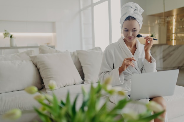 Jovem mulher bonita em roupão de banho fazendo maquiagem diária enquanto assiste a um tutorial em vídeo no laptop