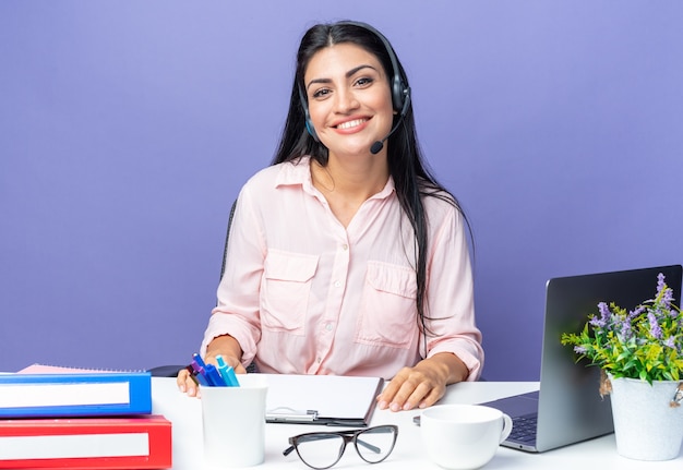 Foto jovem mulher bonita em roupas casuais com fones de ouvido e microfone olhando sorrindo alegremente sentada à mesa com laptop sobre fundo azul trabalhando no escritório