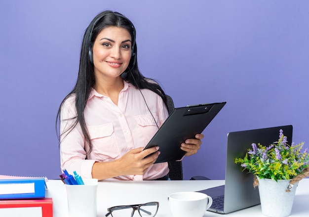 Jovem mulher bonita em roupas casuais, usando fone de ouvido com microfone segurando a prancheta, sorrindo confiante sentada à mesa com o laptop em azul