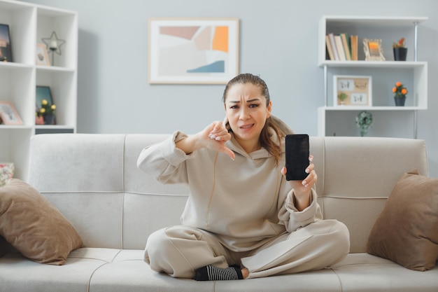 Jovem mulher bonita em roupas de casa sentada em um sofá no interior de casa segurando o smartphone mostrando o polegar para baixo sendo descontente