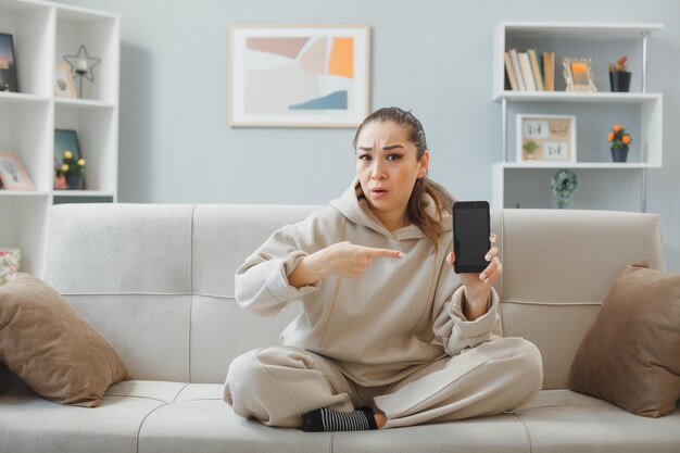 Jovem mulher bonita em roupas para casa, sentado em um sofá no interior de casa, segurando o smartphone apontando com o dedo indicador para ele com expressão cética no rosto, passando o tempo em casa