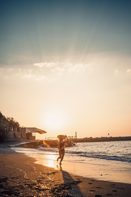 Jovem mulher bonita em um maiô preto e chapéu com óculos caminha ao longo da praia ao pôr do sol O conceito de recreação do mar Foco seletivo