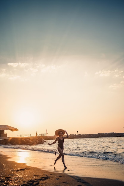 Foto jovem mulher bonita em um maiô preto e chapéu com óculos caminha ao longo da praia ao pôr do sol o conceito de recreação do mar foco seletivo