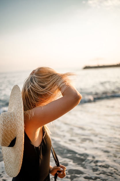 Jovem mulher bonita em um maiô preto e um chapéu com óculos caminha ao longo da praia na Turquia ao pôr do sol. O conceito de recreação marítima. Foco seletivo