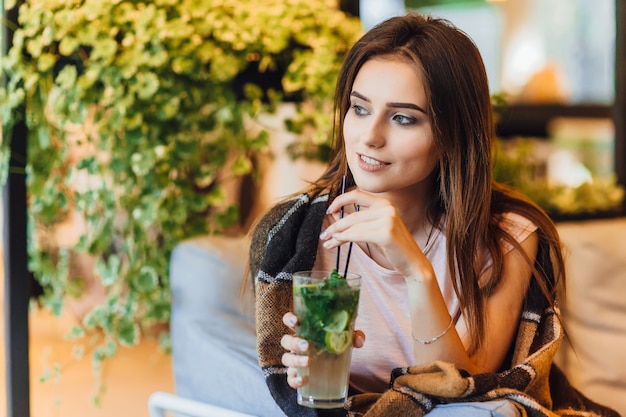 Jovem mulher bonita em um terraço de verão em roupas casuais está bebendo um coquetel.