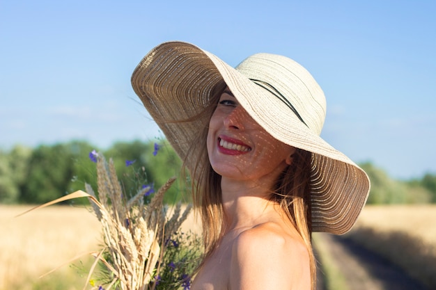 Jovem mulher bonita em um vestido branco e chapéu detém um buquê com flores silvestres na estrada entre o campo leitoso.