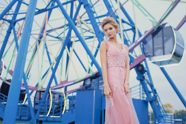 Jovem mulher bonita em um vestido de noite rosa longo caminho no parque. retrato de estilo fashion de linda garota linda ao ar livre perto da roda gigante