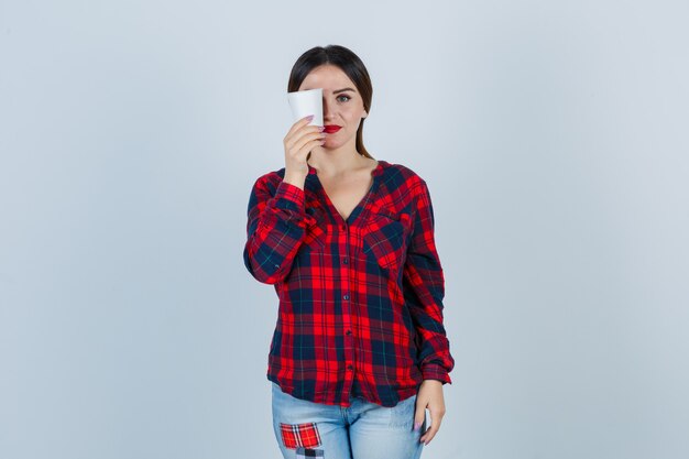 Jovem mulher bonita em uma camisa casual, jeans segurando um copo de plástico no olho e olhando alegre, vista frontal.