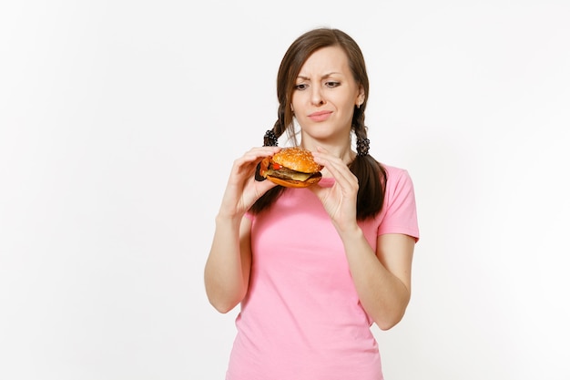 Jovem mulher bonita em uma camiseta rosa com tranças segurando nas mãos hambúrguer isolado no fundo branco. Nutrição adequada ou fast food clássico americano. Copie o espaço para anúncio. Área de publicidade.