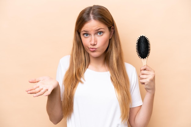 Jovem mulher bonita loira com pente de cabelo isolado em fundo bege fazendo gesto de dúvidas ao levantar os ombros