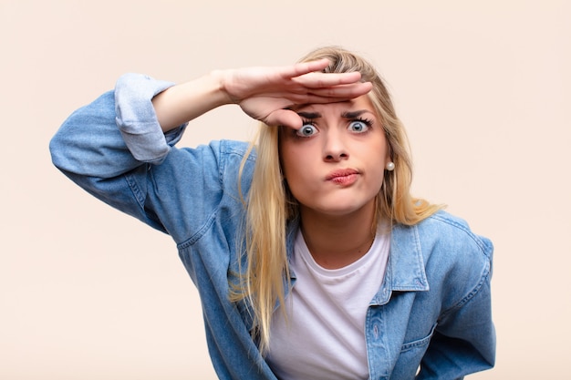 Foto jovem mulher bonita loira olhando confuso e surpreso, com a mão na testa, olhando para longe, assistindo ou pesquisando contra parede plana