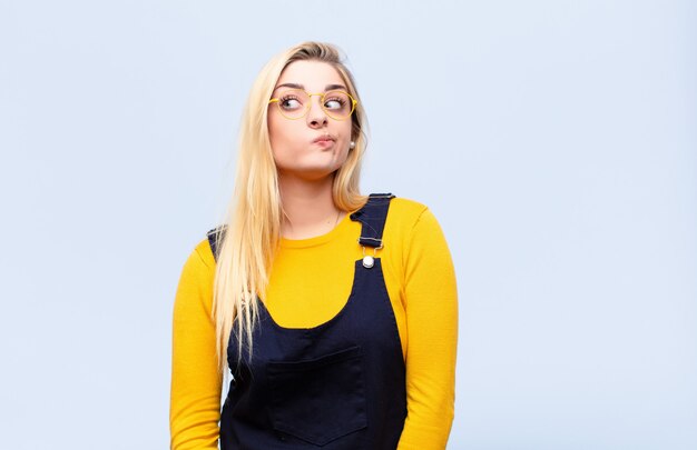 Jovem mulher bonita loira pensando, pensando felizes pensamentos e idéias, sonhando acordado, olhando para copiar o espaço do lado contra a parede plana