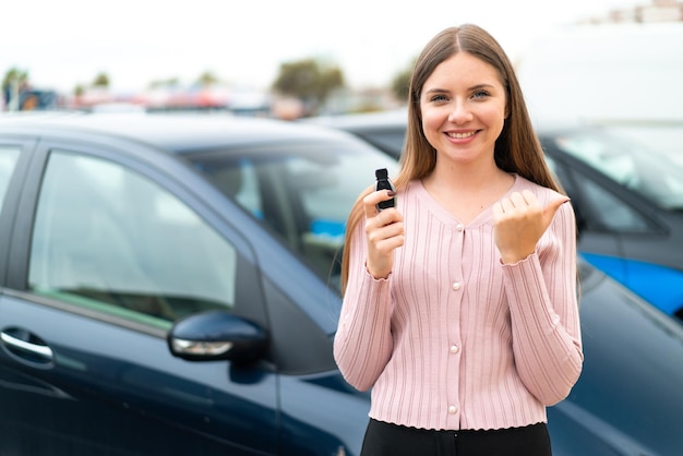 Jovem mulher bonita loira segurando as chaves do carro ao ar livre apontando para o lado para apresentar um produto