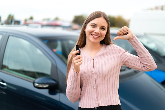 Jovem mulher bonita loira segurando as chaves do carro ao ar livre orgulhosa e satisfeita