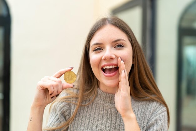 Jovem mulher bonita loira segurando um Bitcoin ao ar livre gritando com a boca aberta