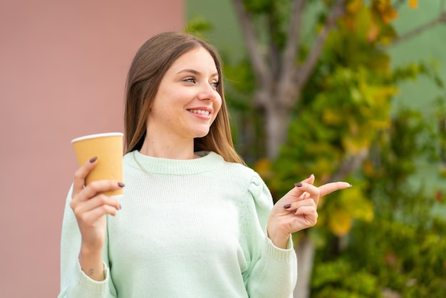 Jovem mulher bonita loira segurando um café para viagem ao ar livre apontando para o lado para apresentar um produto