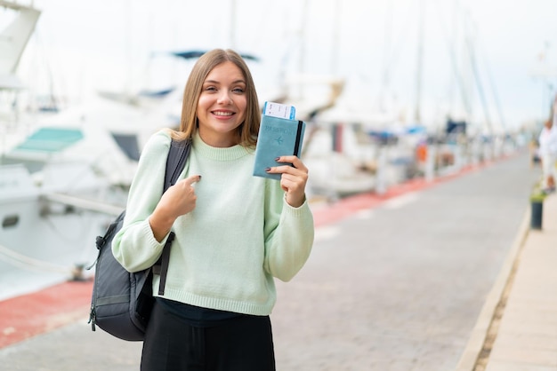 Jovem mulher bonita loira segurando um passaporte ao ar livre com expressão facial de surpresa