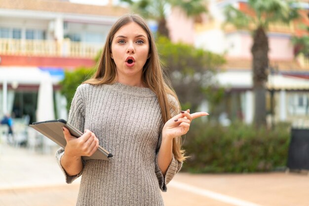 Jovem mulher bonita loira segurando um tablet ao ar livre surpreso e apontando para o lado