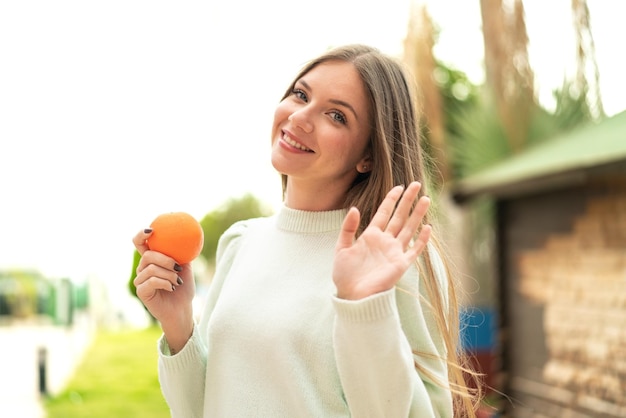 Jovem mulher bonita loira segurando uma laranja ao ar livre saudando com a mão com expressão feliz